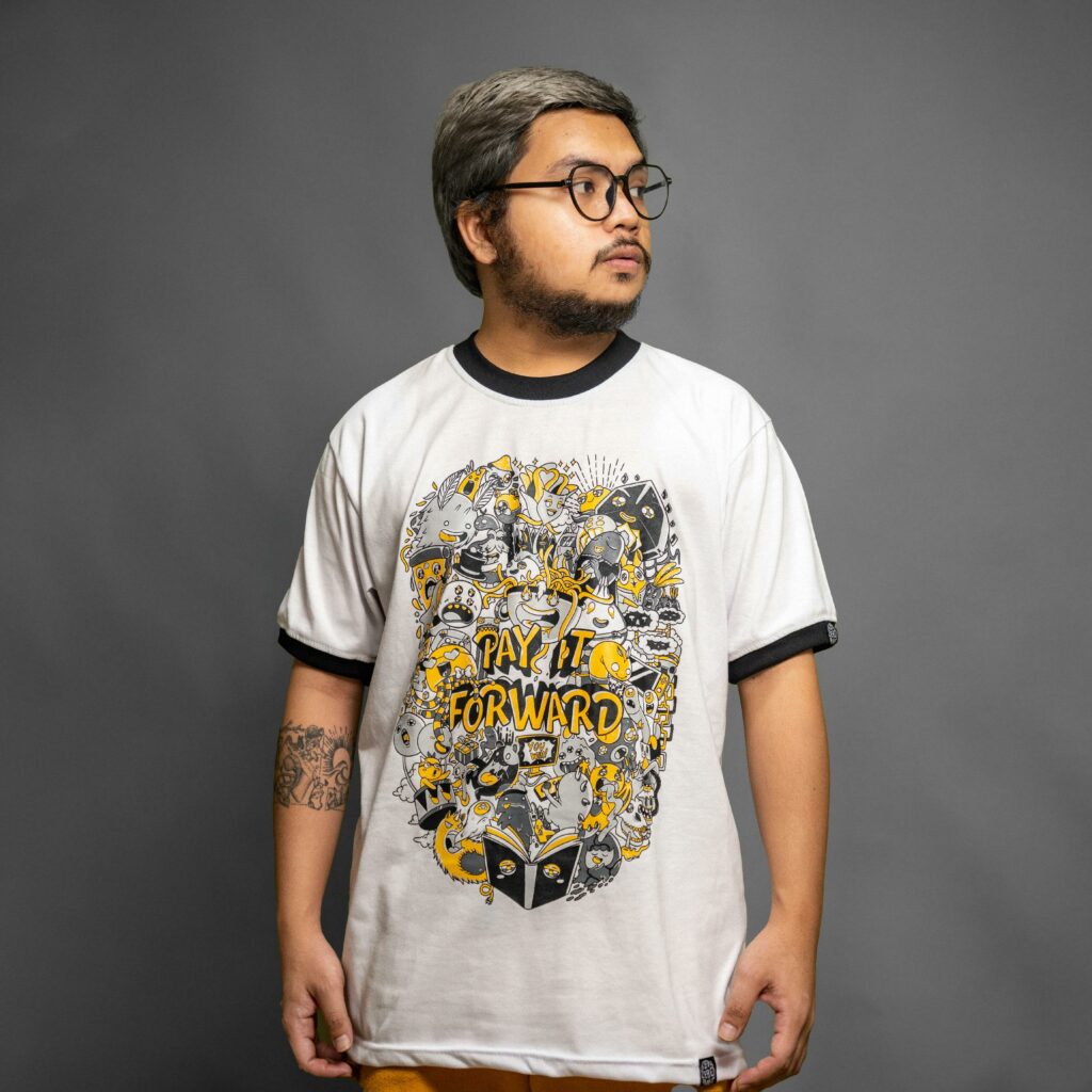 Bearded man in a white 'Pay It Forward' t-shirt posing in a studio setting.